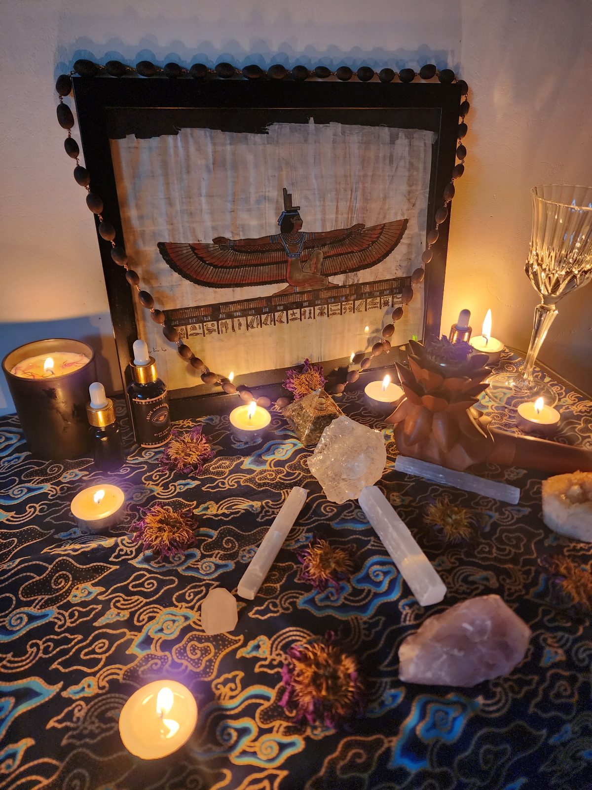 A home altar with a papyrus painting of a winged goddess figure, candles, and crystals, prepared for Blue Lotus Initiation work.