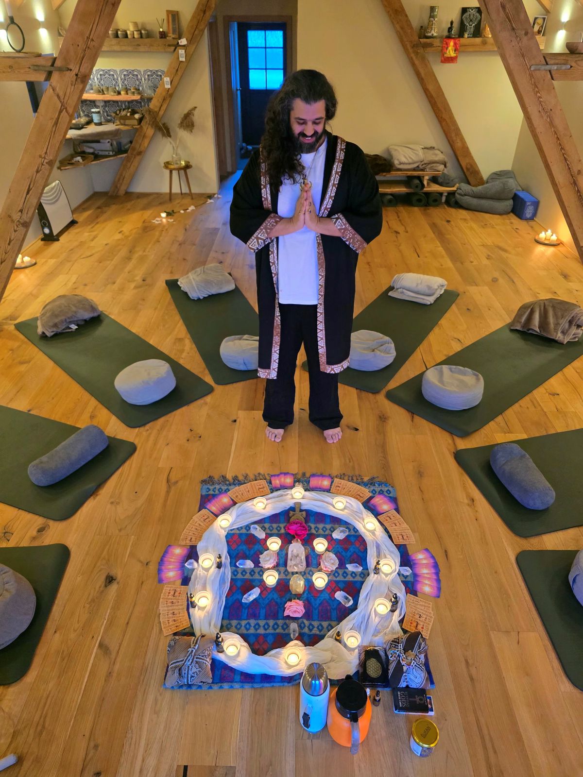 Ali Odeh standing in a ceremony room, mats and cushions set in a circle around a lit altar.