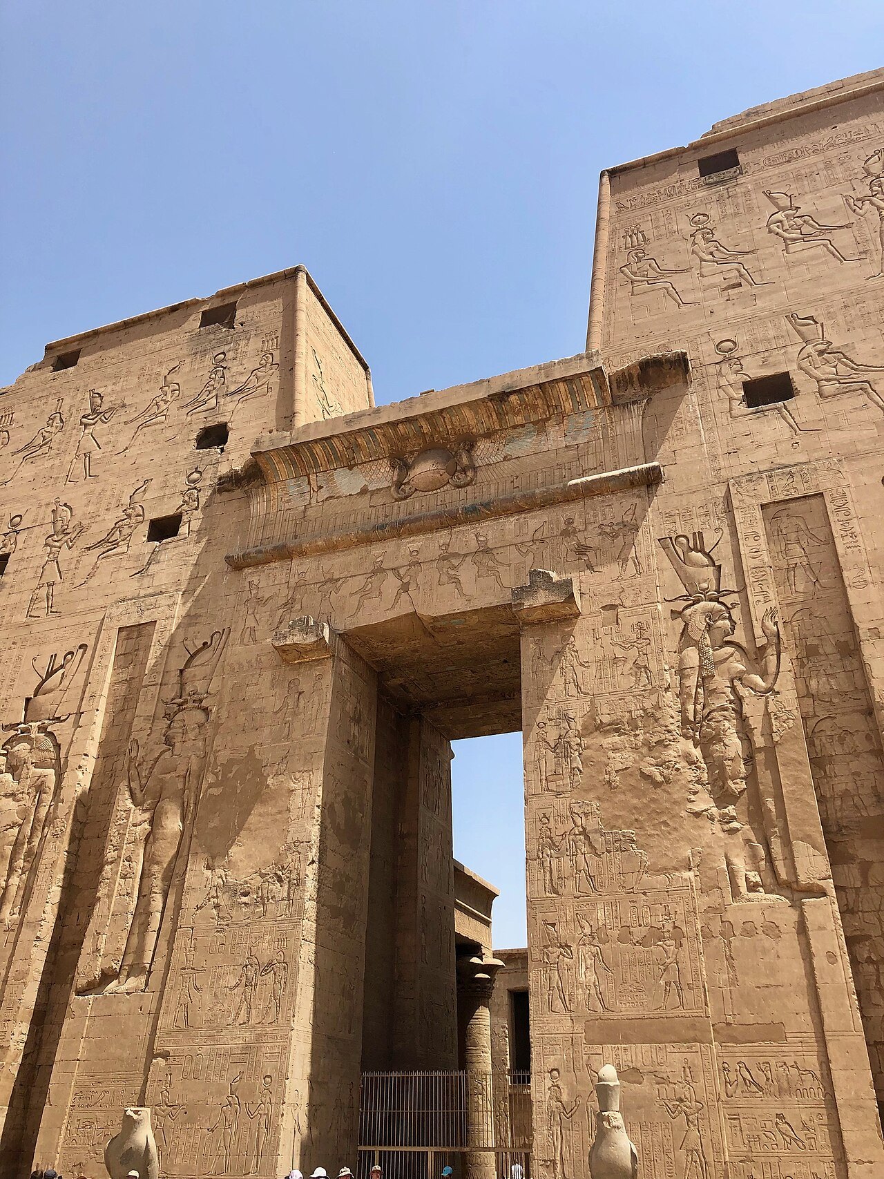 The great pylon of the Temple of Horus at Edfu, its towers carved with figures, the gateway open against a blue sky.